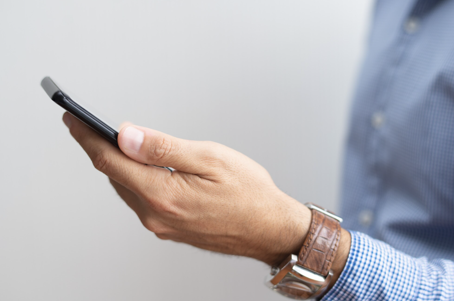 Man's hand holding black smartphone, brown leather watch on wrist.